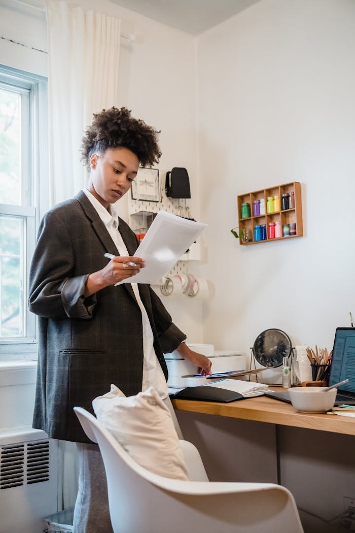 Young woman in home office reviewing documents by the desk with tech setup.
